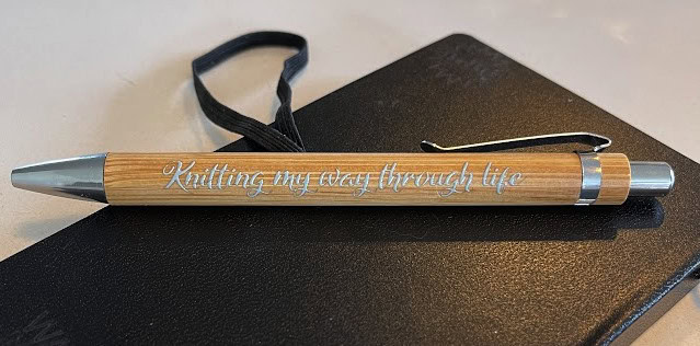 Custom wooden pen printed with "Knitting my every through life" on a black notebook.